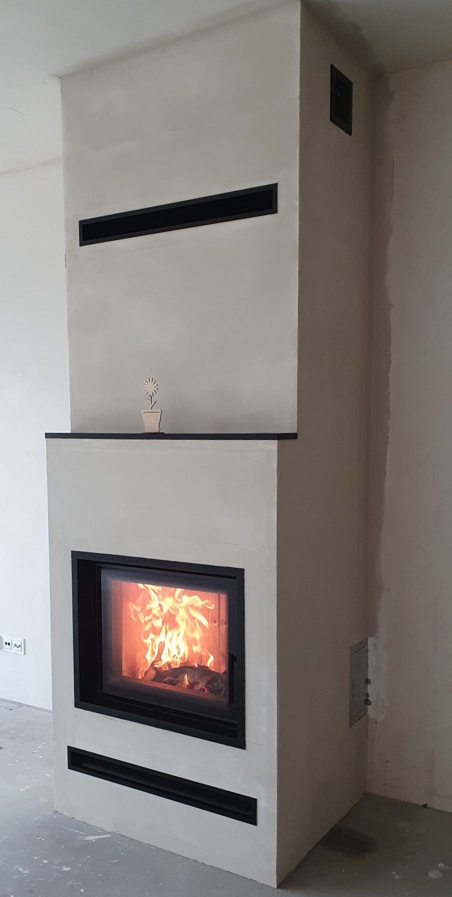 Nowoczesny kominek z czarną obudową i płomieniami, ozdobiony drewnianą dekoracją kwiatową na półce, z widocznymi kratkami wentylacyjnymi.