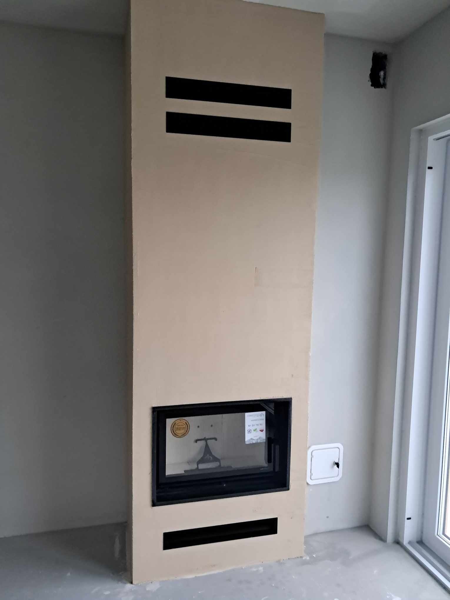 Zamknięty kominek z beżową obudową i czarnymi akcentami wentylacyjnymi w surowym wnętrzu, widok z przodu, w tle ściana i okno.