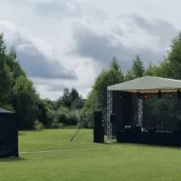 Scena plenerowa z nagłośnieniem i oświetleniem, rozstawiona na zielonym trawniku, obok ciemny namiot, drzewa w tle, pochmurne niebo.