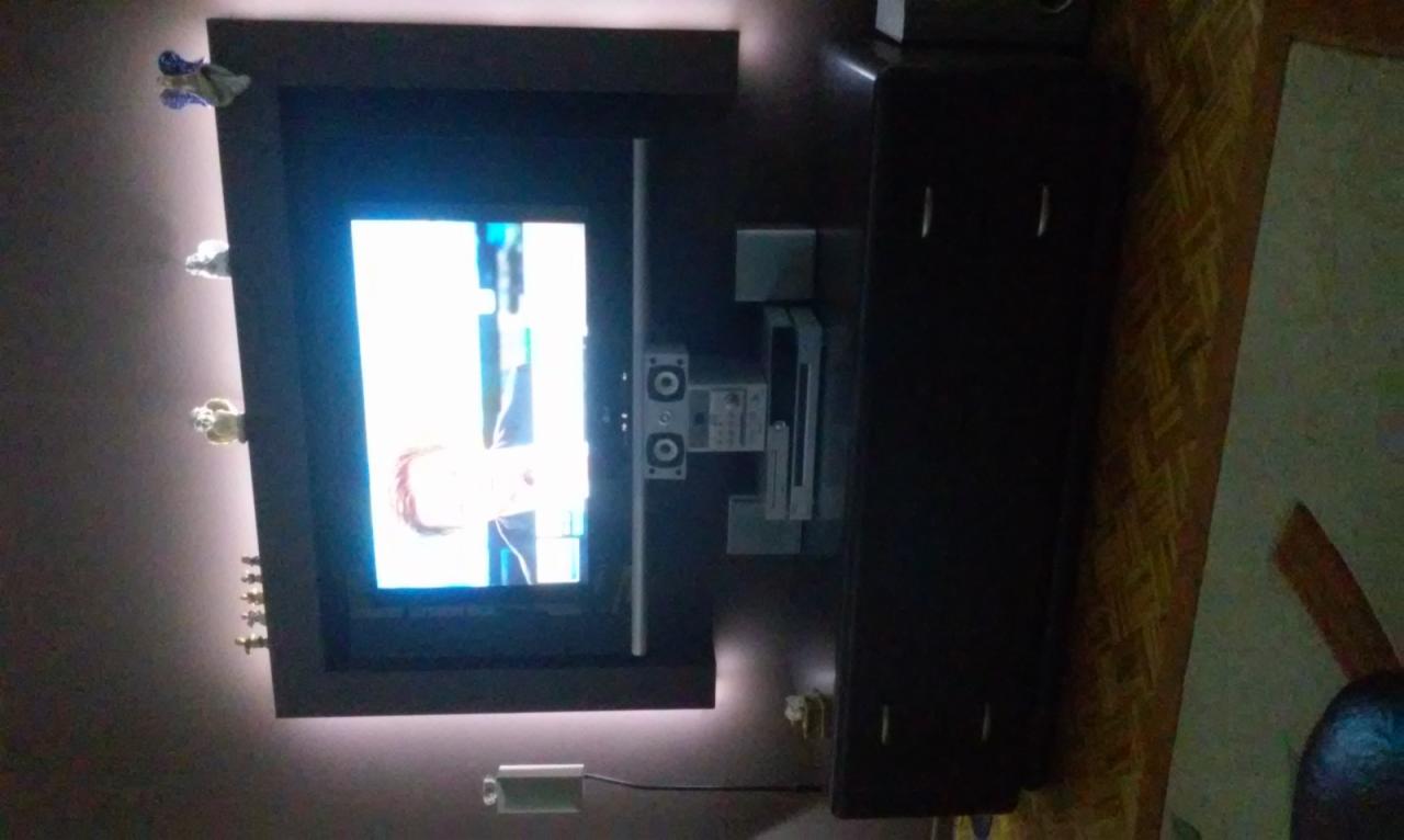 Telewizor w ciemnej obudowie z podświetleniem LED, ozdobiony figurkami na ścianie, obok szafka RTV. Kątowe ujęcie.