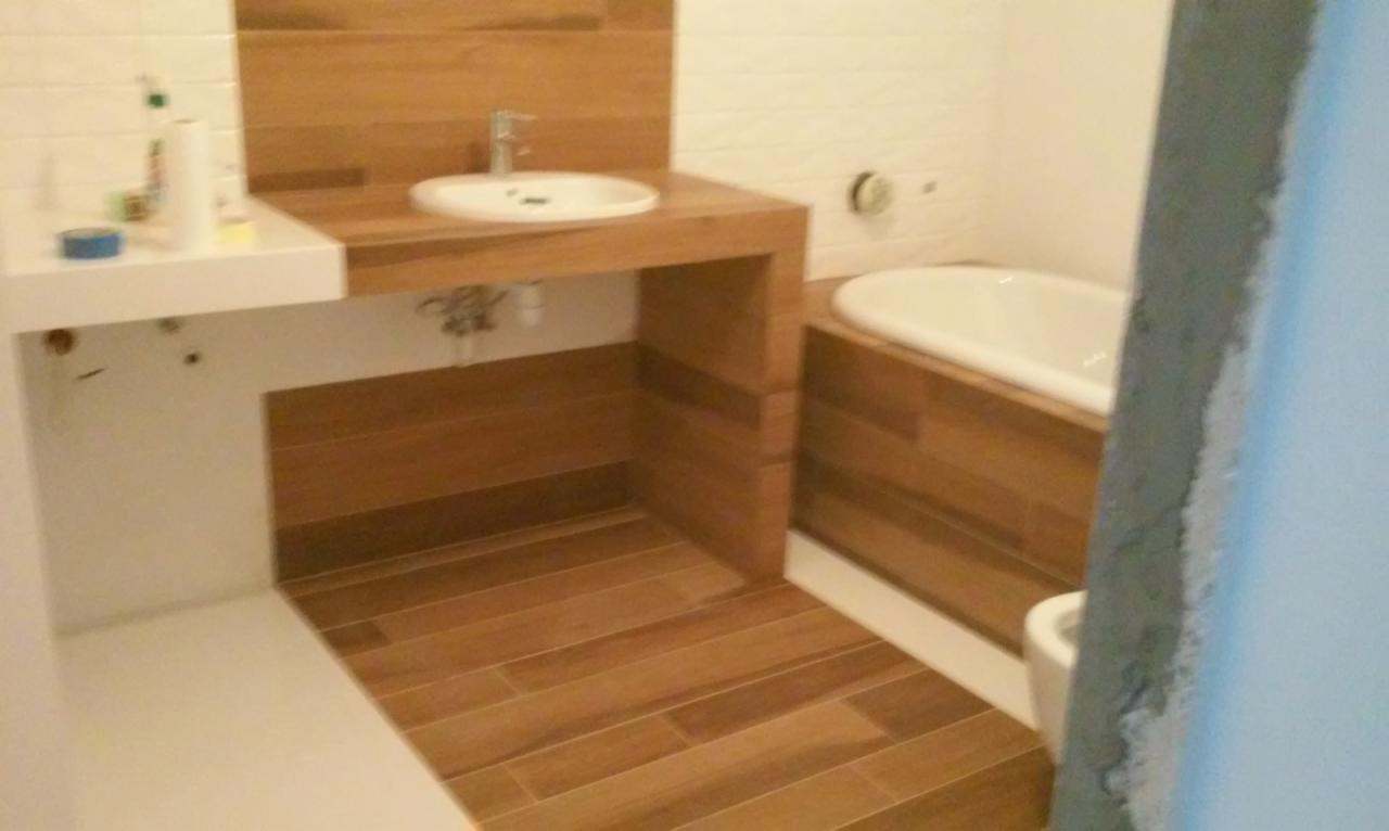 Nowoczesna łazienka po remoncie: drewnopodobne płytki, zabudowa wanny i umywalki, biała armatura. Widok z perspektywy wejścia, z surowym fragmentem ściany.