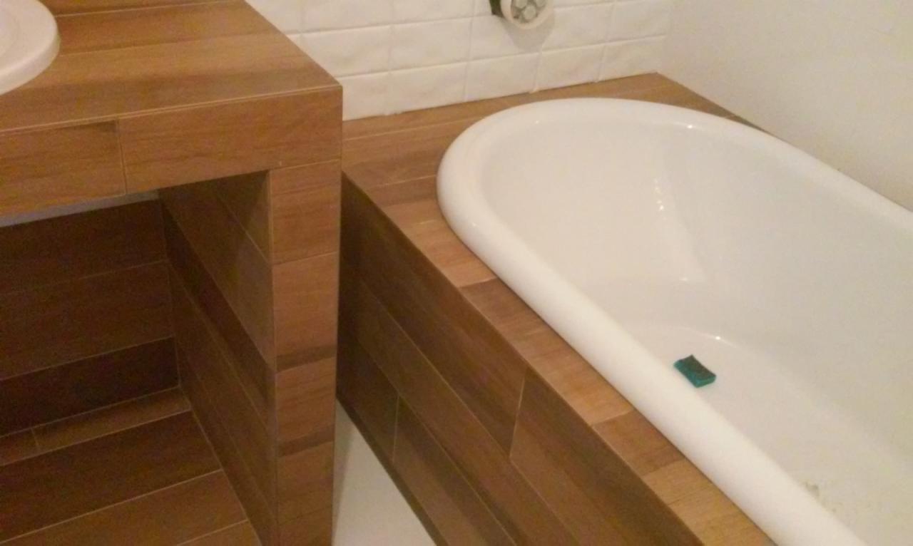 Wykończona łazienka: biała wanna z obudową z drewnopodobnych płytek, dopasowana zabudowa umywalki, białe płytki na ścianie. W wannie leży gąbka.