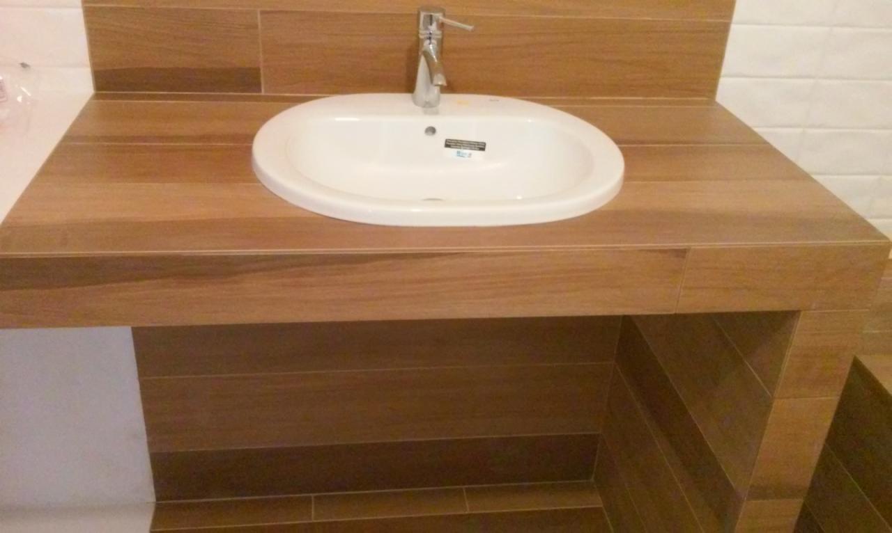 Umywalka wpuszczona w drewnopodobny blat z płytek, bateria chromowana, biała cegiełka na ścianie. Aranżacja łazienki z bliska.