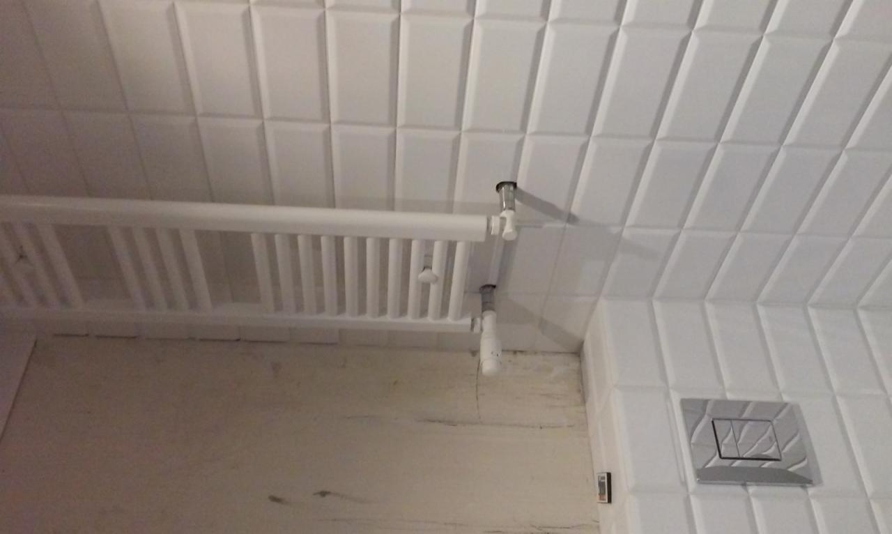 Biały grzejnik łazienkowy na ścianie z białych, błyszczących płytek. Widoczne rury doprowadzające wodę i przycisk spłuczki. Ściana w trakcie wykończenia.