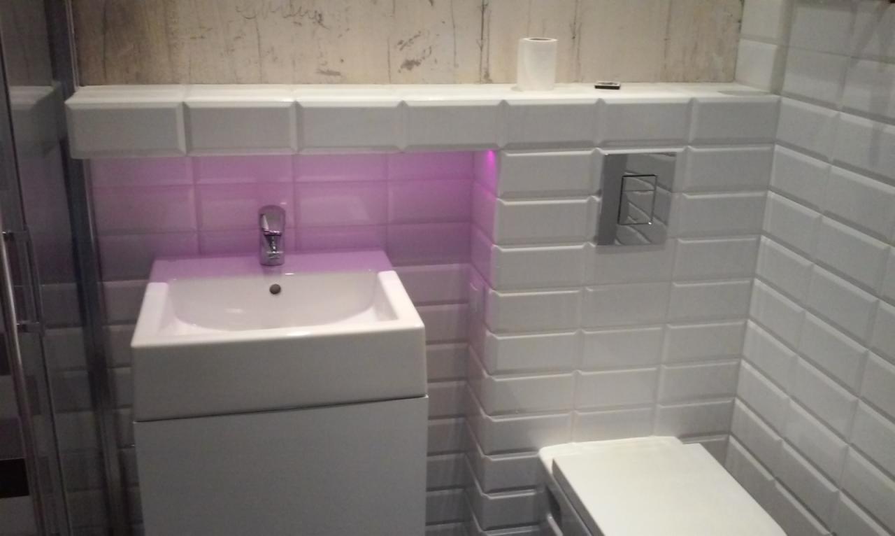 Biała łazienka z umywalką, toaletą i podświetlanymi płytkami. Umywalka z szafką, bateria umywalkowa. Płytki ułożone w cegiełkę. Nowoczesny design łazienki.