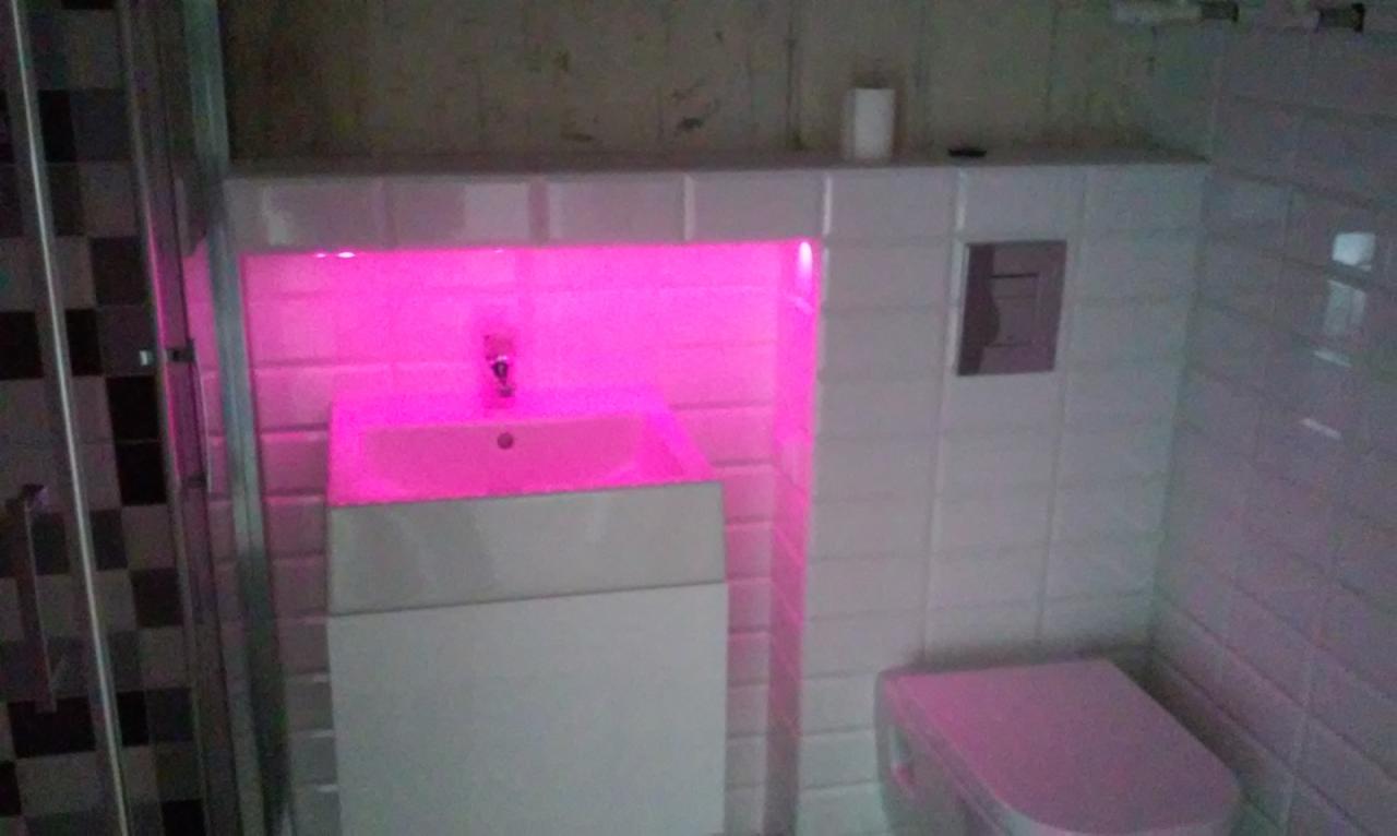 Wykończona łazienka z podświetlaną wnęką nad umywalką, białe płytki i nowoczesna toaleta w odcieniach różu. Widoczny fragment kabiny prysznicowej.