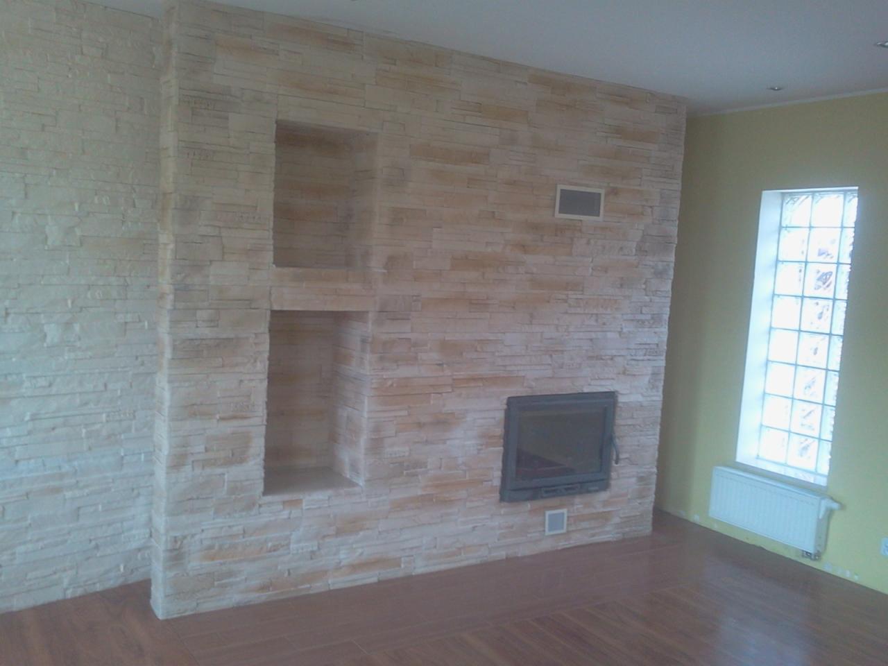 Wykończona ściana z kamienia dekoracyjnego z wnękami i kominkiem, podłoga z paneli, okno z luksferów, widok na fragment pokoju po remoncie.