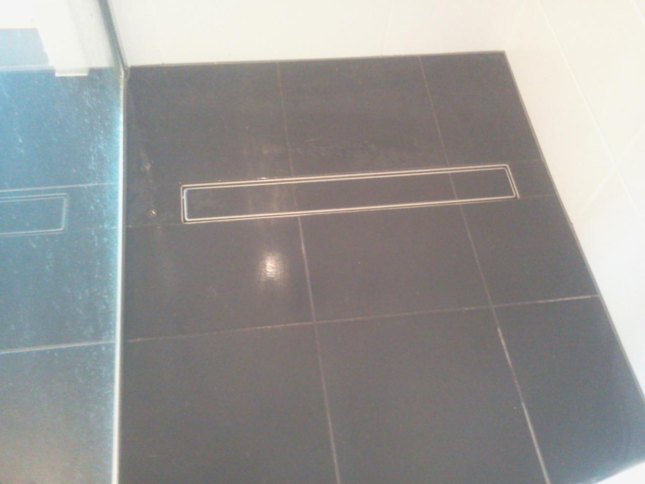 Ciemne płytki podłogowe w łazience z odpływem liniowym. Widoczna kabina prysznicowa z boku. Minimalistyczny design, proste linie, nowoczesne wykończenie.