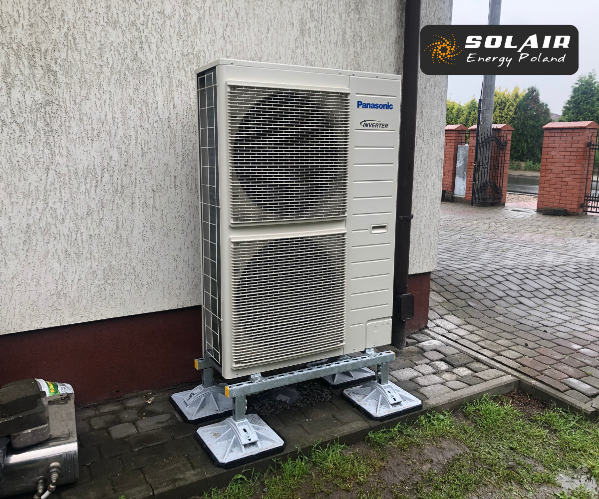Zewnętrzna jednostka pompy ciepła Panasonic Inverter zamontowana na metalowej konstrukcji przy ścianie budynku z widocznym logo Solair Energy Poland w prawym górnym rogu.
