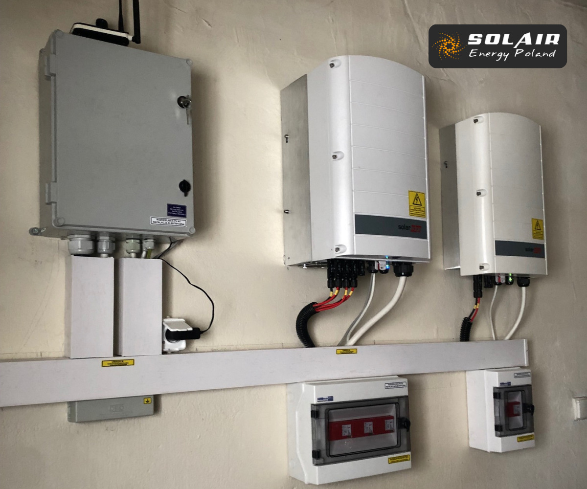 Szara skrzynka elektryczna z anteną, dwa białe inwertery z żółtymi naklejkami ostrzegawczymi, przewody i bezpieczniki zamontowane na ścianie wewnątrz budynku.