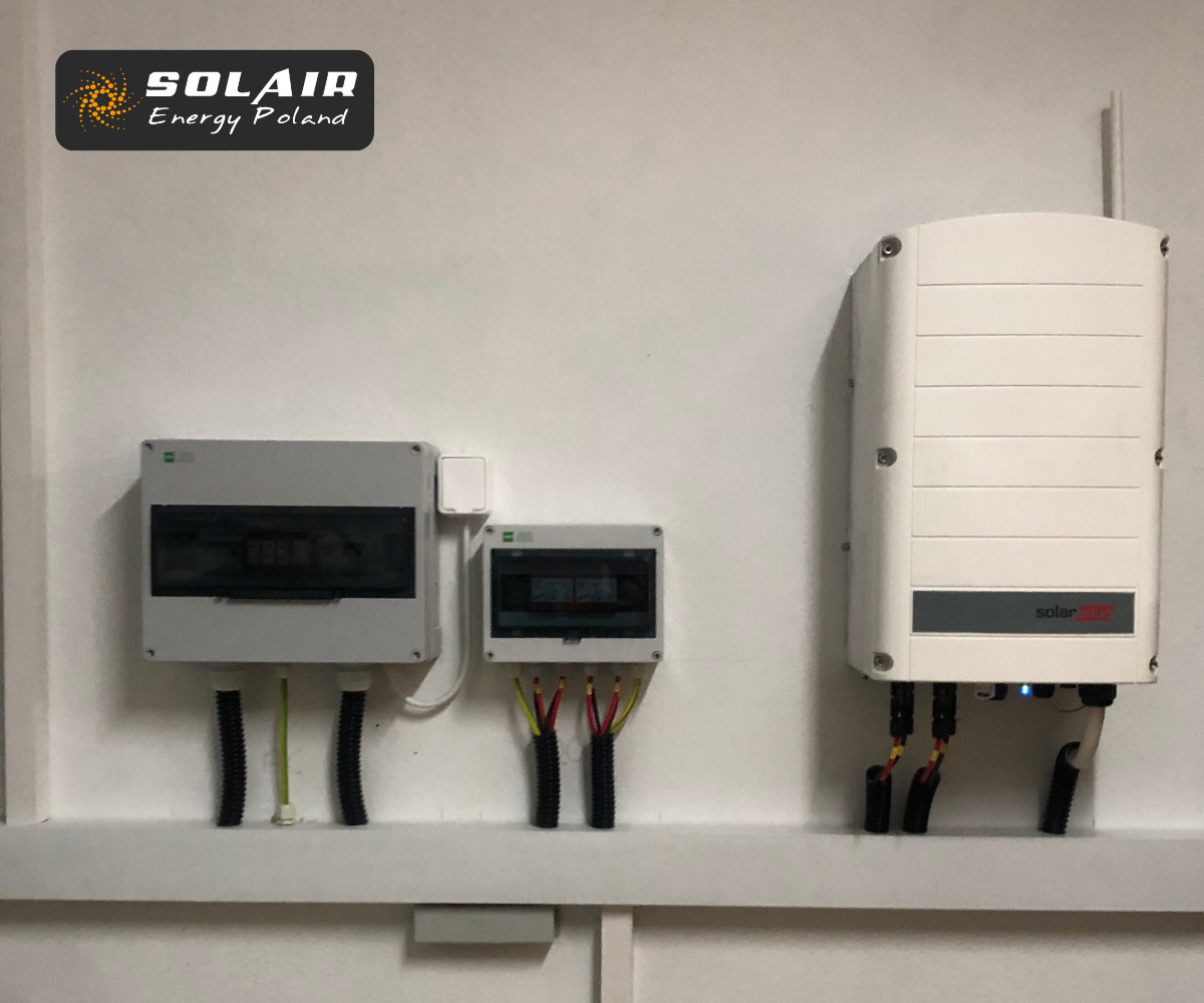 Biała ściana z dwoma szarymi skrzynkami rozdzielczymi i białym falownikiem z anteną, widoczne przewody w peszlach, logo firmy w lewym górnym rogu.