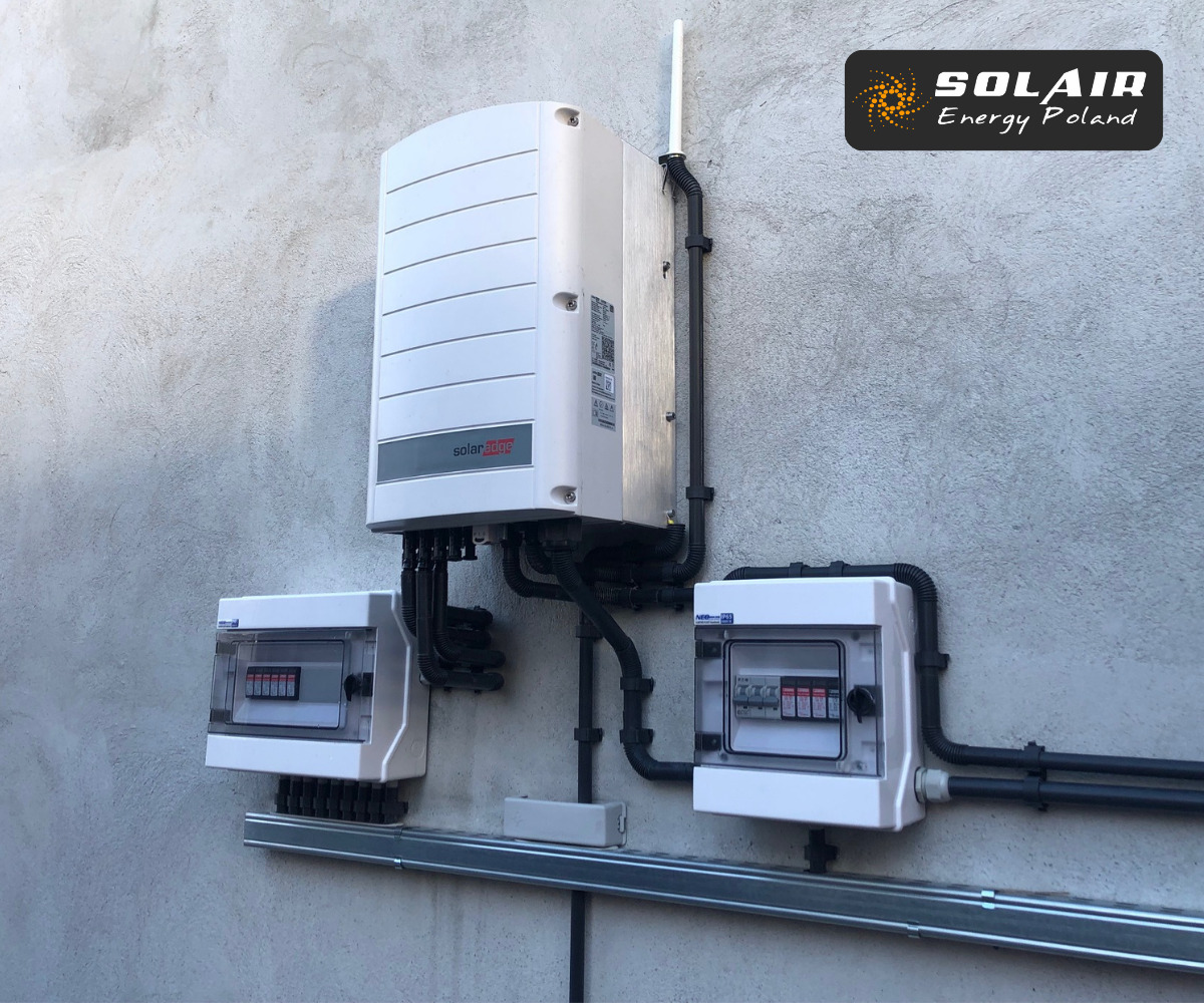 Biały inwerter SolarEdge zamontowany na szarej, betonowej ścianie, z dwoma skrzynkami bezpiecznikowymi poniżej, przymocowanymi do metalowej szyny.