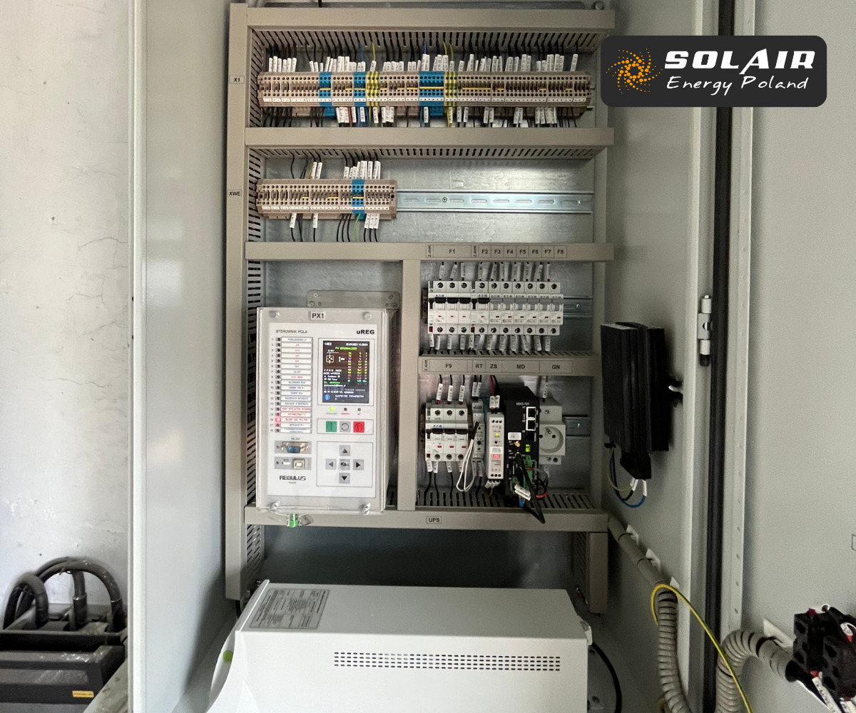 Otwarta szafa sterownicza z aparaturą elektryczną, widoczne zabezpieczenia, sterownik z wyświetlaczem oraz moduły z okablowaniem, dolna część szafy zajęta przez biały zasilacz UPS.