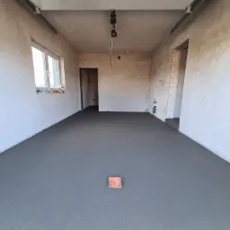 Świeżo wylana, gładka, szara podłoga w surowym pomieszczeniu z widocznymi instalacjami elektrycznymi i hydraulicznymi w ścianach. Na środku podłogi leży pomarańczowa płytka dystansowa.