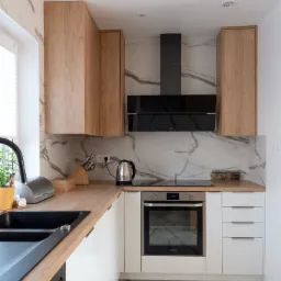 Nowoczesna kuchnia z marmurową ścianą, drewnianymi szafkami wiszącymi i czarnym zlewem, oświetlona światłem dziennym.