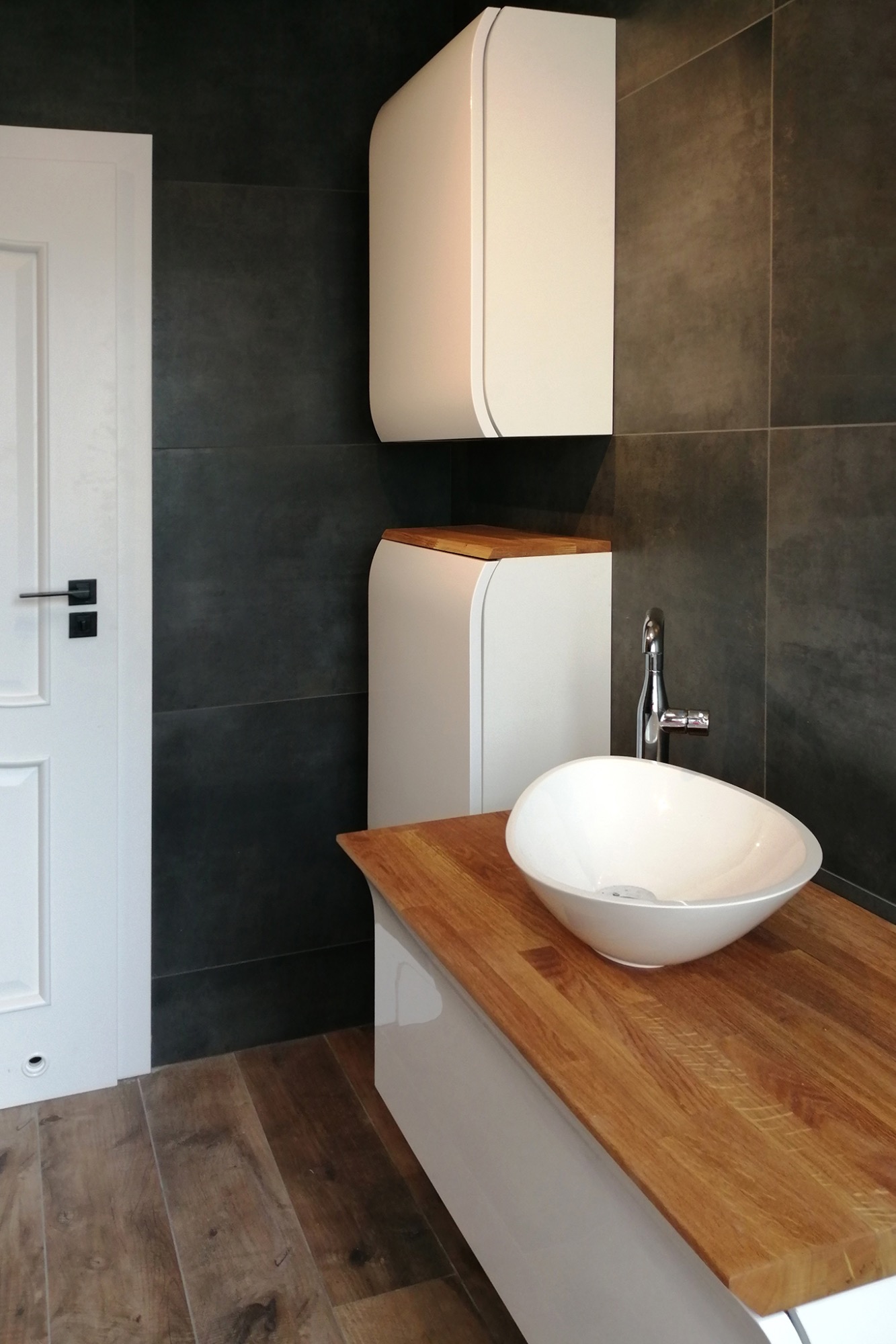 Nowoczesna łazienka z umywalką nablatową w kształcie misy, drewnianym blatem, białymi szafkami o zaokrąglonych kształtach i ciemnoszarą, kwadratową glazurą na ścianach.