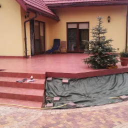 Wykończony taras z brązowych płytek ceramicznych z widocznymi schodami, na których leżą narzędzia glazurnicze, oraz przygotowanym podłożem pod przyszły ogród, obok rośnie mała choinka.