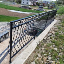 Czarna, metalowa balustrada z dekoracyjnymi elementami, oddzielająca taras z kostki brukowej od skarpy z kamieniami i trawą, w tle widoczny krajobraz wiejski z domami i polami.