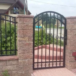 Czarna, metalowa brama wjazdowa z ozdobnymi elementami, osadzona na murowanych słupkach z piaskowca, z widocznym napędem elektrycznym, uzupełniona o ogrodzenie z przęsłami o podobnym wzorze, na tle...