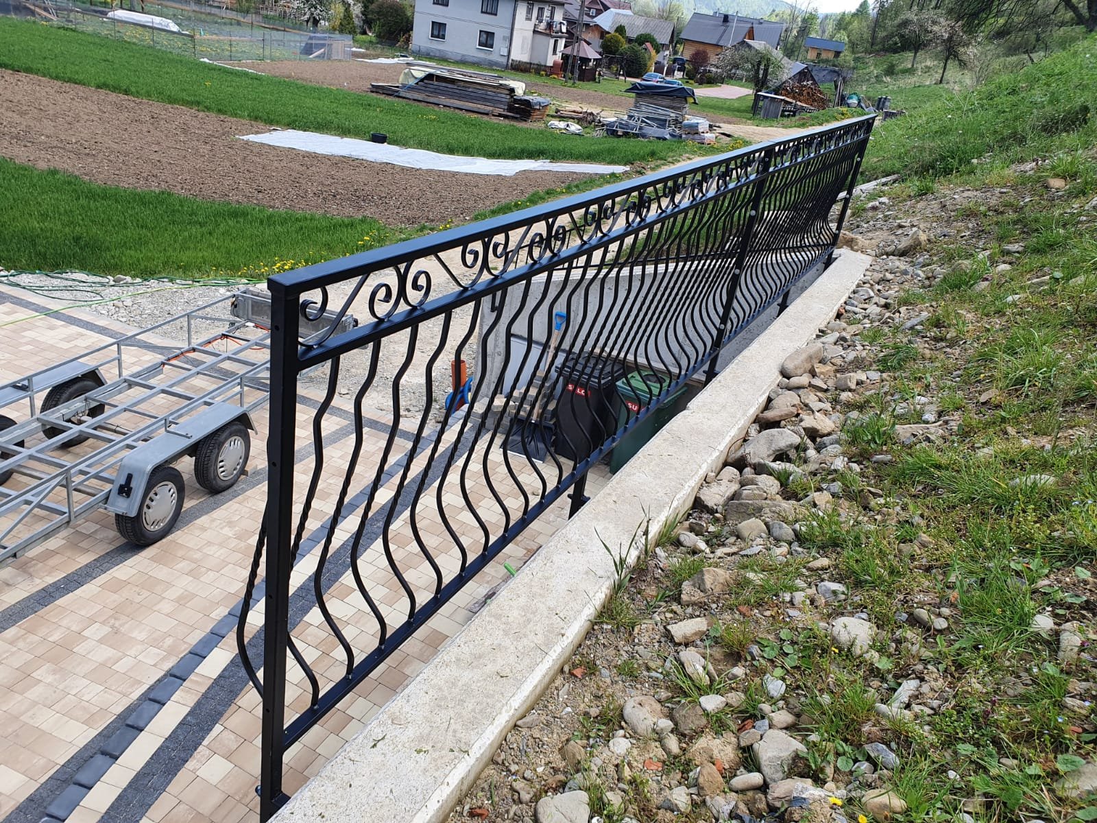 Czarna, metalowa balustrada z dekoracyjnymi elementami, oddzielająca taras z kostki brukowej od skarpy z kamieniami i trawą, w tle widoczny krajobraz wiejski z domami i polami.