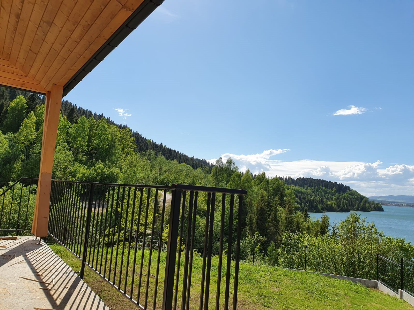 Czarna metalowa balustrada na tarasie z widokiem na zalesione wzgórza i jezioro pod błękitnym niebem z chmurami.