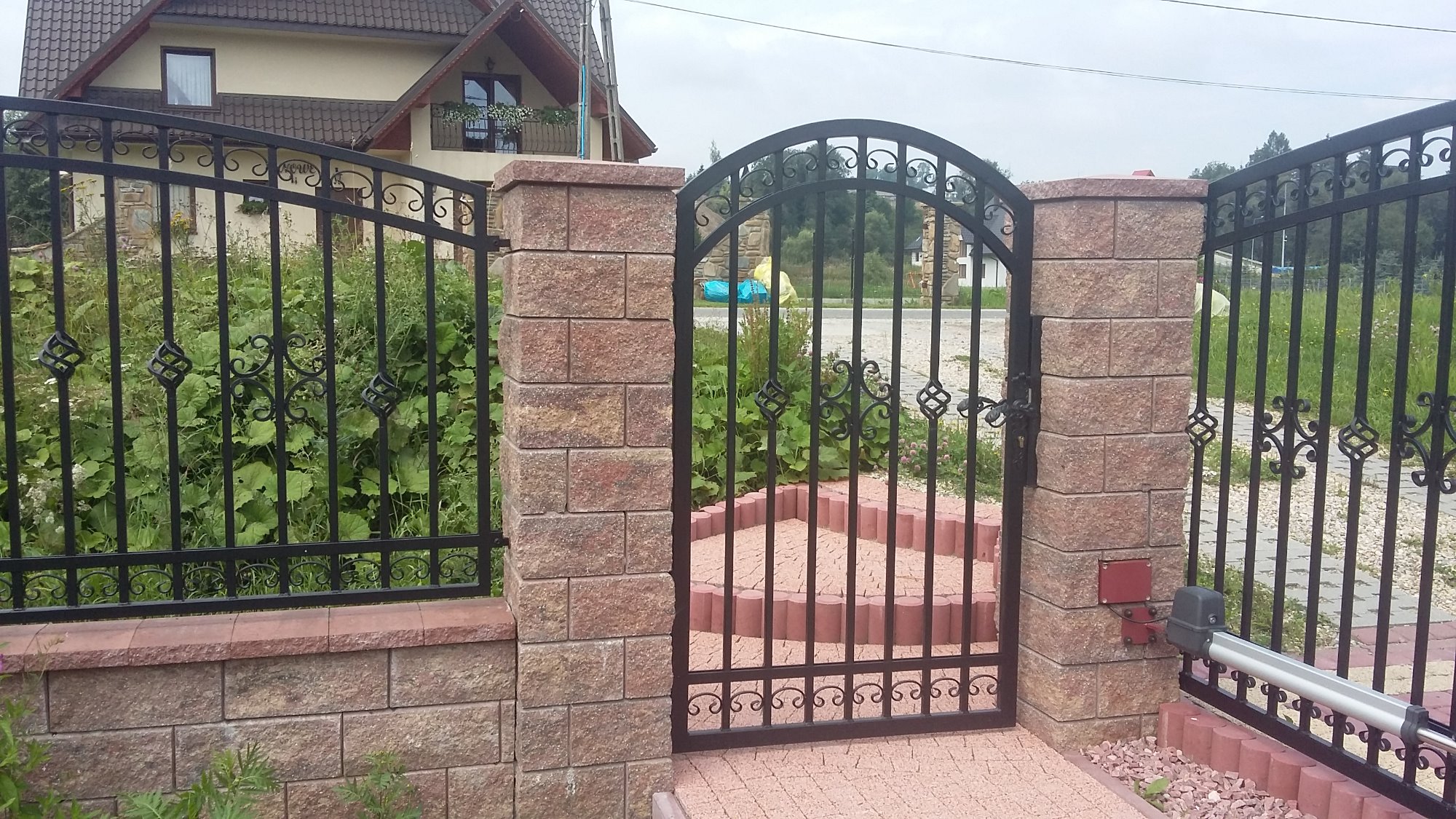 Czarna, metalowa brama wjazdowa z ozdobnymi elementami, osadzona na murowanych słupkach z piaskowca, z widocznym napędem elektrycznym, uzupełniona o ogrodzenie z przęsłami o podobnym wzorze, na tle...