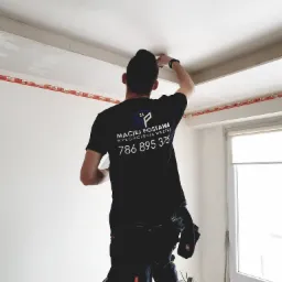 Mężczyzna na drabinie wyrównuje szpachlą łączenie płyt kartonowo-gipsowych na suficie, widoczna taśma wzmacniająca, na koszulce logo firmy i numer telefonu.