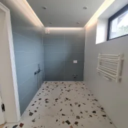 Nowoczesna łazienka z szarymi płytkami ściennymi, podświetleniem LED pod sufitem i terazzo na podłodze, grzejnik drabinkowy na ścianie.