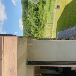 Surowy stan elewacji budynku z widocznym ociepleniem, fragmentem drewnianej boazerii, kostką brukową i zielenią w tle.