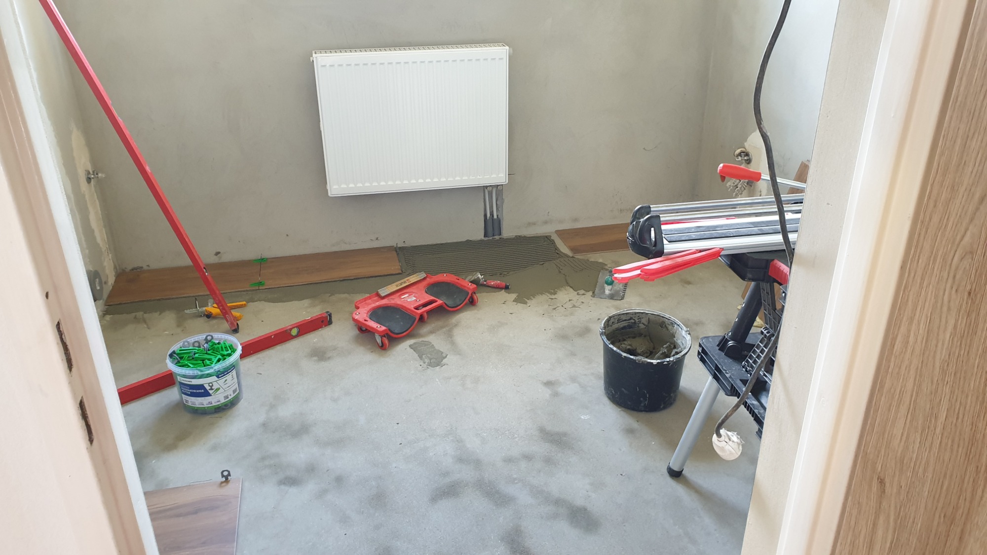 Przygotowanie do układania drewnopodobnych płytek w łazience, widoczny rozprowadzony klej, poziomica, wiadro z zaprawą, przyssawka do przenoszenia płytek oraz maszyna do cięcia płytek, ogrzewanie...