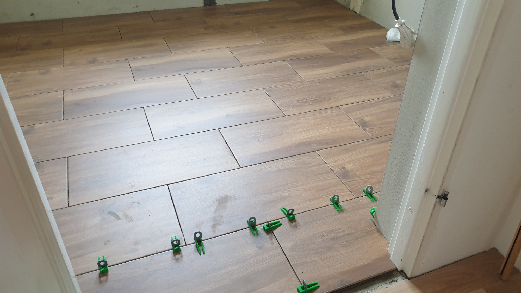 Układanie podłogowych płytek imitujących drewno z użyciem zielonych klipsów poziomujących przy białej futrynie drzwi.