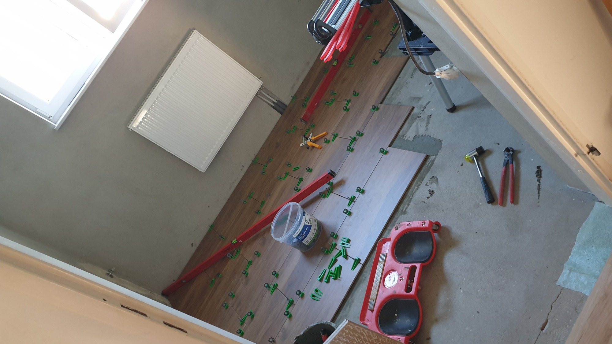 Układanie brązowych płytek podłogowych w łazience z użyciem poziomicy, klipsów poziomujących i wiadra z zaprawą, widoczne narzędzia glazurnicze oraz grzejnik na ścianie.