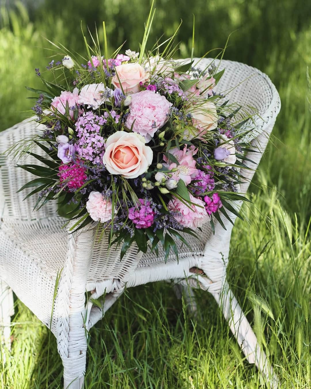 Bukiet różowych i fioletowych kwiatów, w tym piwonii i róż, ułożony na białym, wiklinowym krześle na tle zielonej trawy w Warszawie.