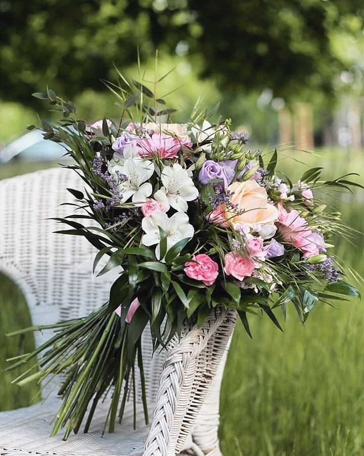 Delikatny bukiet z róż, alstromerii i lawendy, oparty o białe, wiklinowe krzesło na tle zielonego trawnika i drzew.