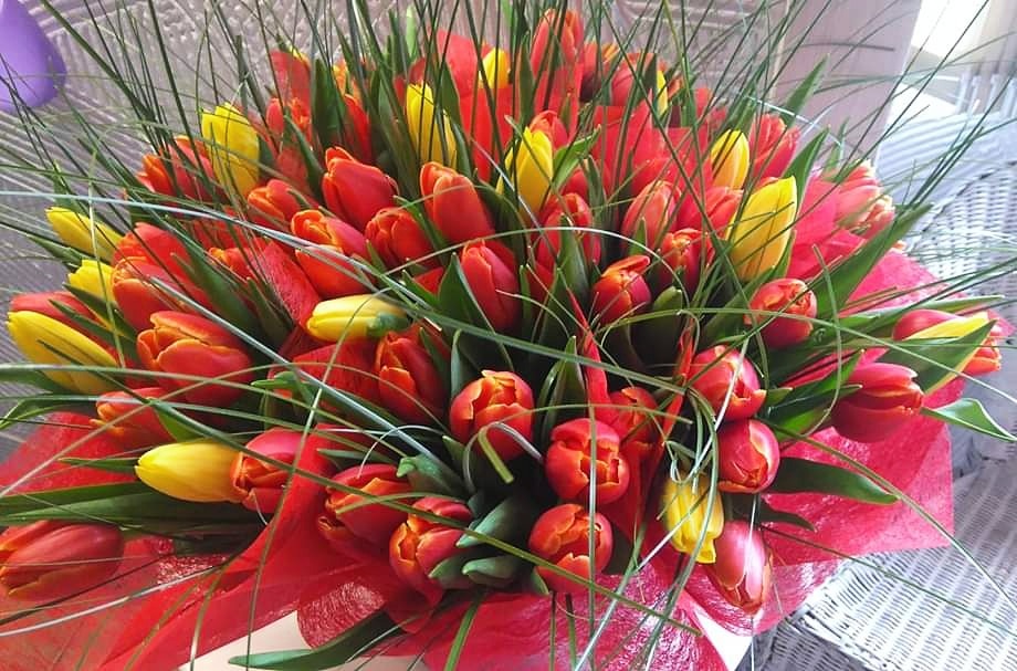 Gęsty bukiet czerwono-żółtych tulipanów, owinięty czerwoną, połyskującą folią, ozdobiony długimi, zielonymi źdźbłami trawy, ustawiony na białym, plecionym podłożu.
