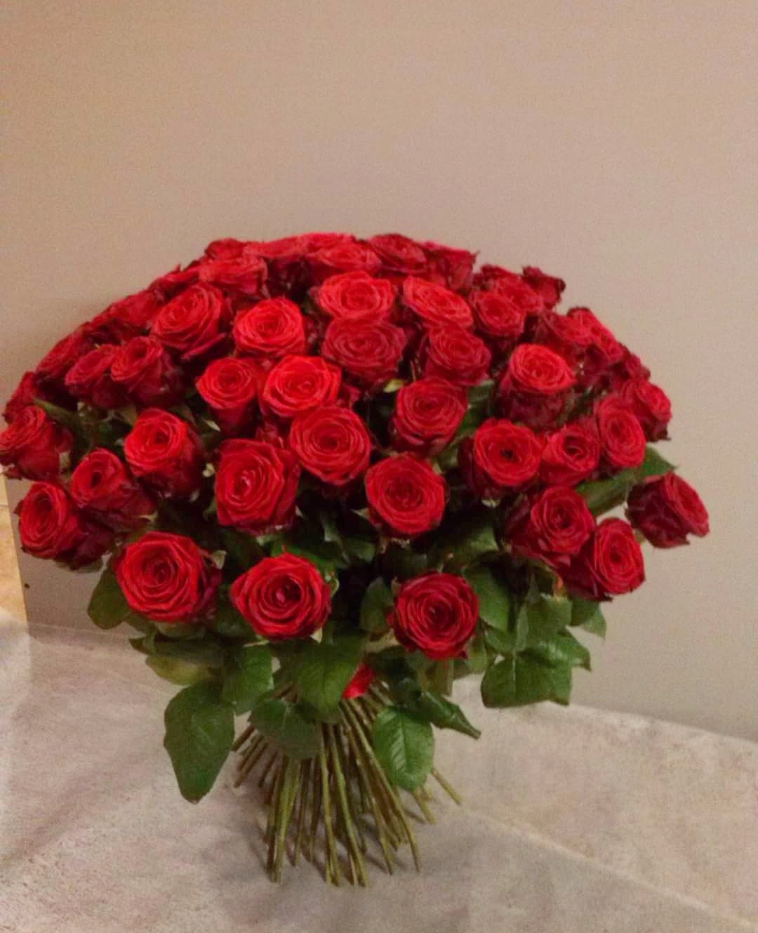 Duży bukiet intensywnie czerwonych róż ułożonych w półkulę, z widocznymi zielonymi liśćmi i łodygami związanymi czerwoną wstążką.