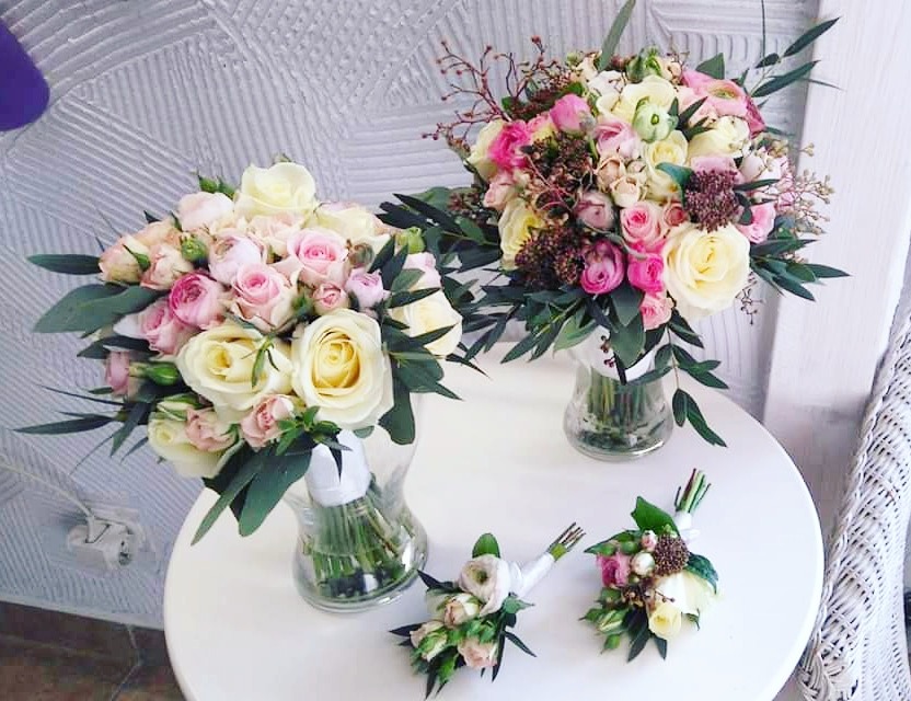 Dwa bukiety z białych i różowych róż w szklanych wazonach na białym okrągłym stole, uzupełnione zielonymi liśćmi eukaliptusa i drobnymi brązowymi dodatkami, wraz z dwoma butonierkami z identycznych...