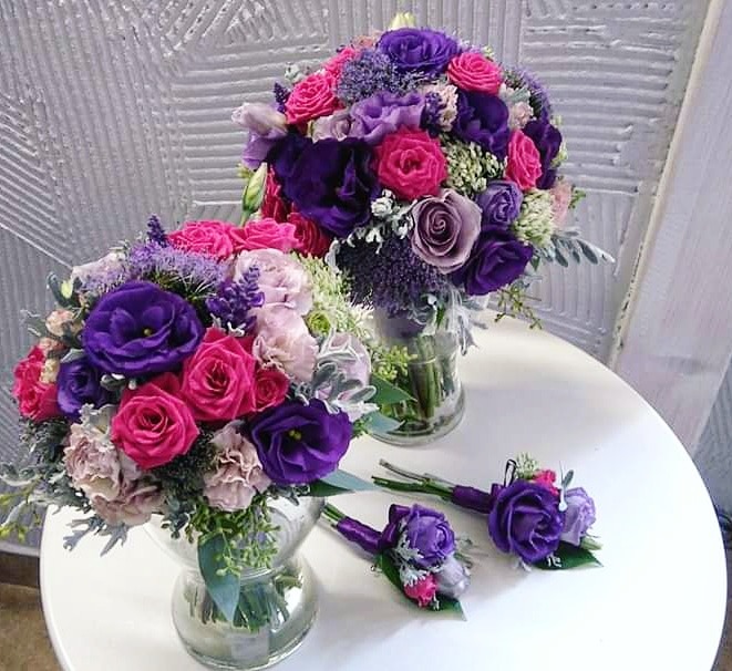 Dwa bukiety z różowych i fioletowych róż w szklanych wazonach oraz dwie butonierki, ułożone na białym okrągłym stole na tle teksturowanej ściany.