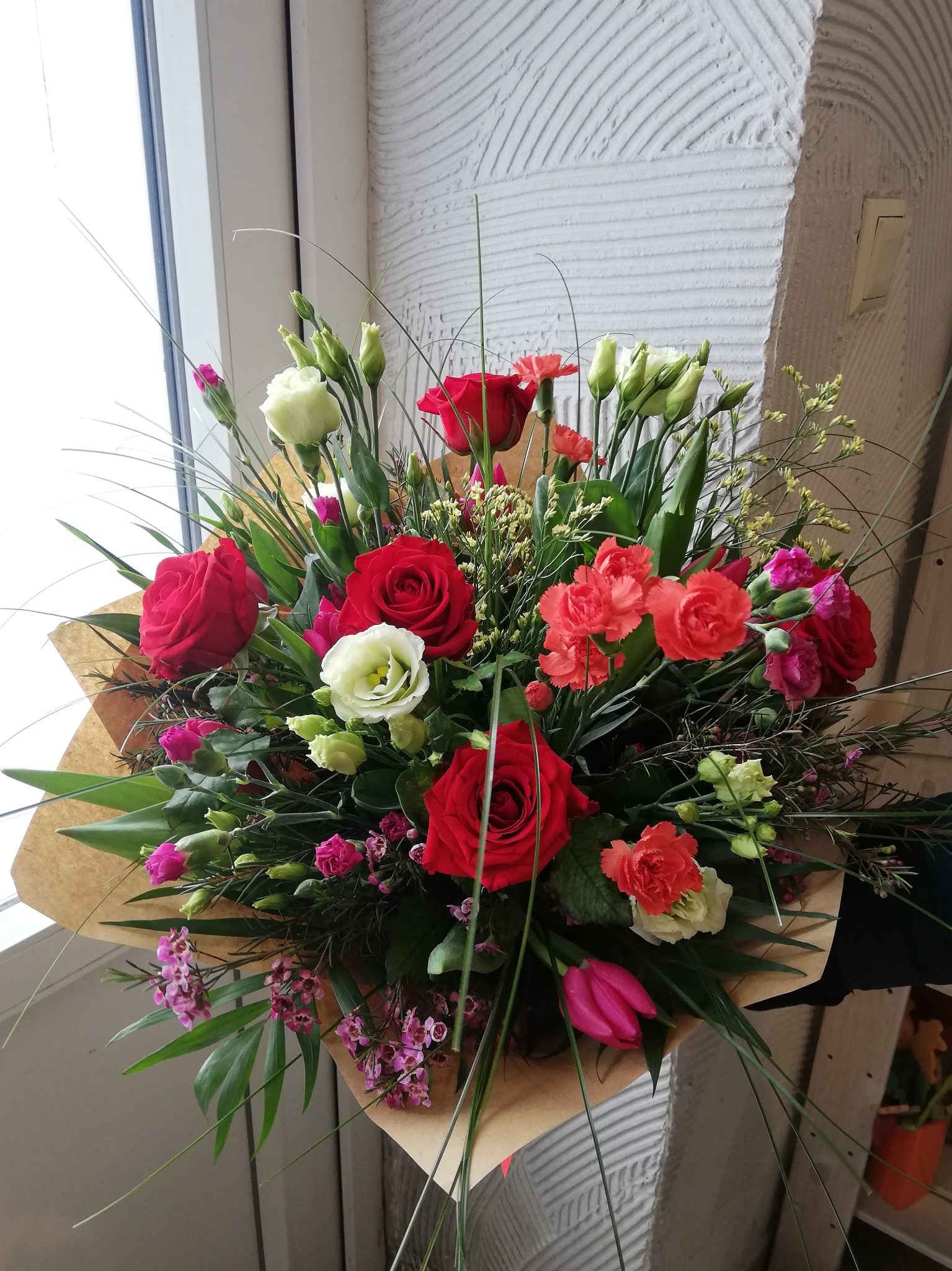 Bogaty bukiet mieszany z czerwonych róż, goździków i białych eustom w brązowym papierze, ustawiony przy oknie z teksturowanym tłem ściany.