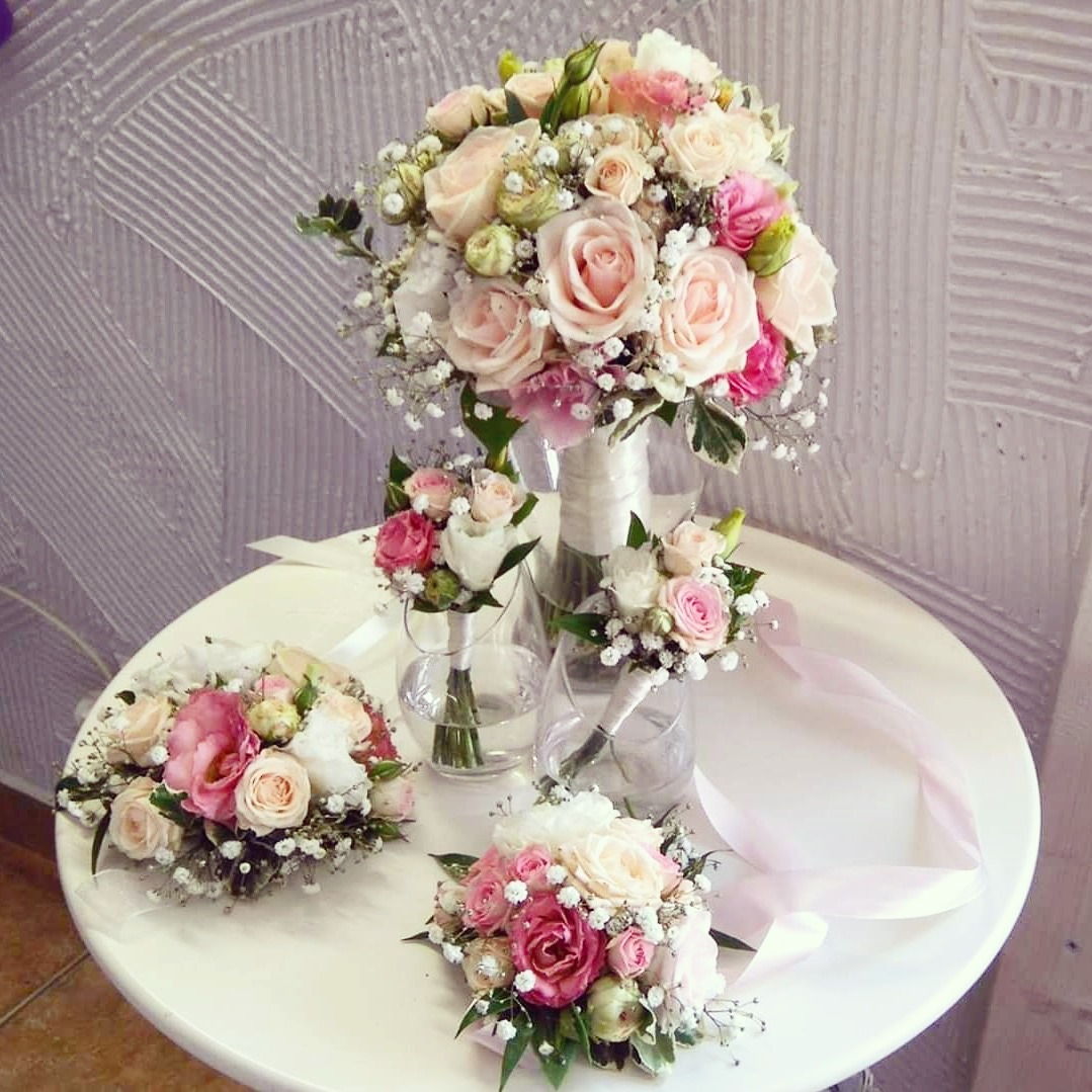 Kompozycja z bukietu ślubnego z róż w odcieniach różu i kremu, oraz mniejszych bukiecików w szklanych wazonach na białym okrągłym stole, z delikatną wstążką w tle.