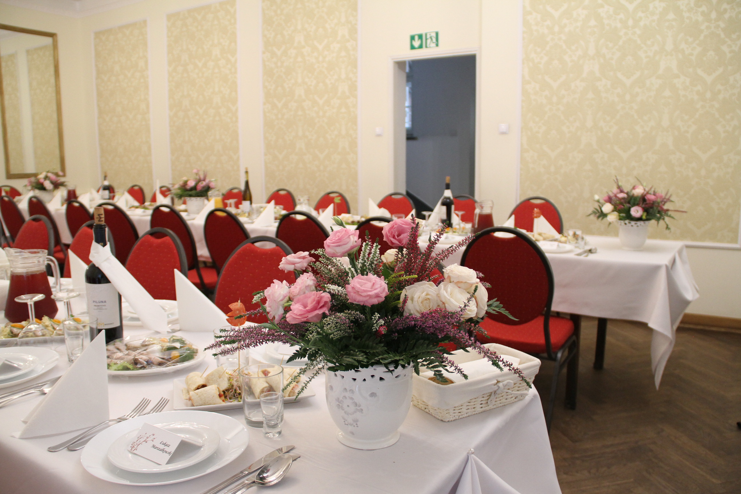 Uroczysta sala bankietowa z nakrytymi stołami, dekoracjami kwiatowymi z róż w białych wazonach, zastawą stołową, napojami i przekąskami.