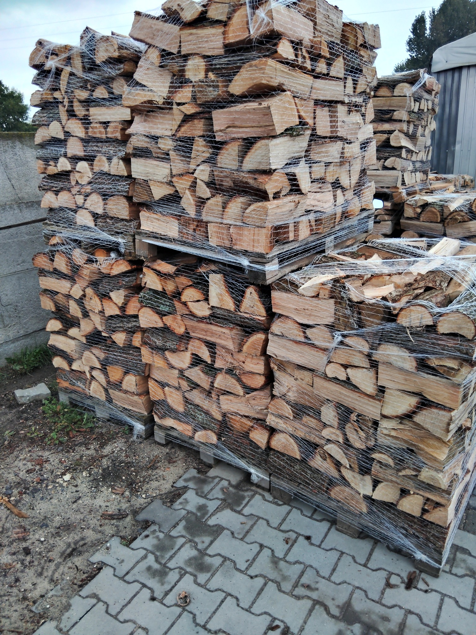 Ułożone na paletach i owinięte siatką pakiety pociętego drewna, składowane na zewnątrz na kostce brukowej.