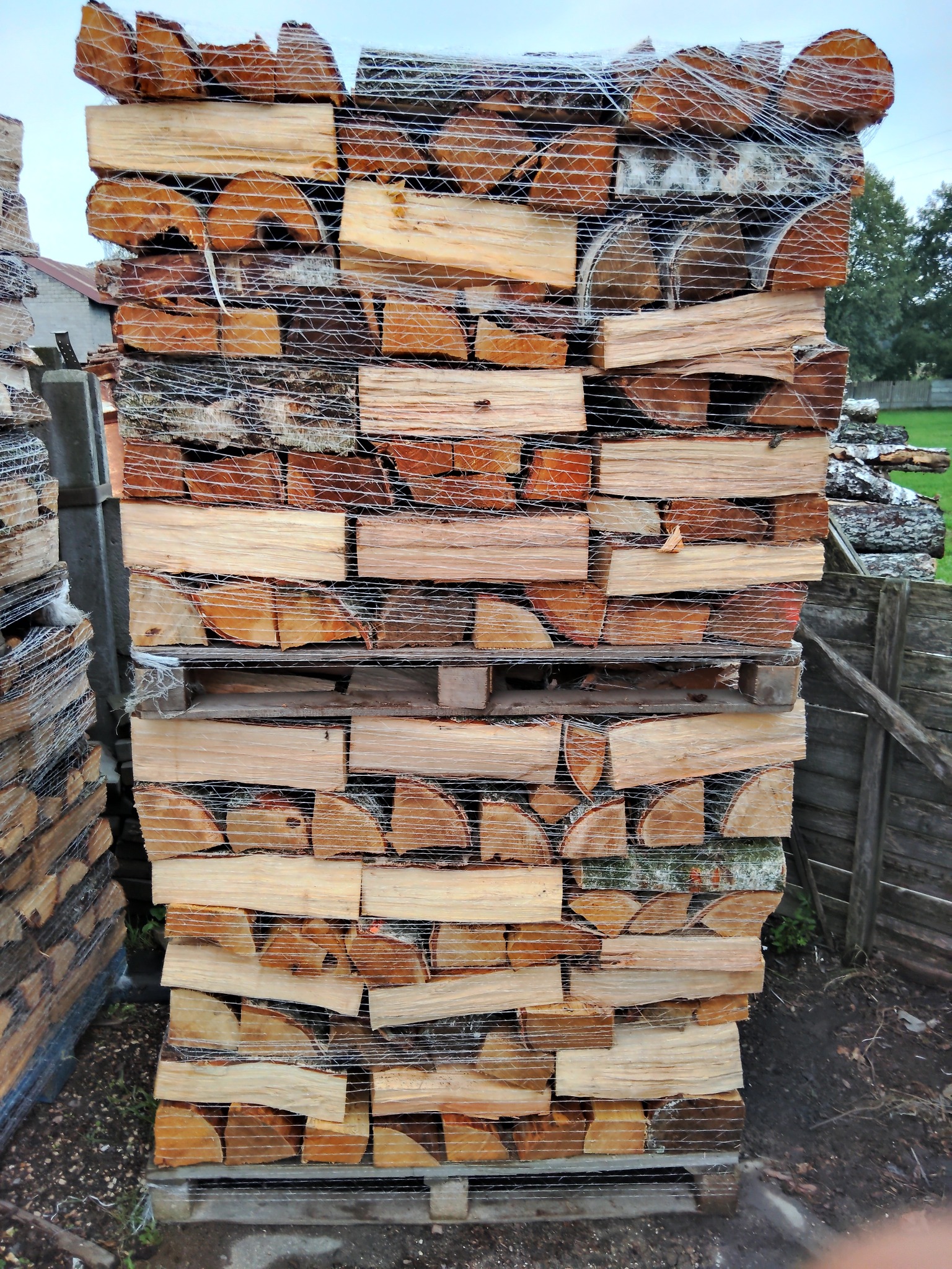 Paleta z ułożonymi rzędami pociętego drewna opałowego, zabezpieczonego siatką, widoczne inne palety w tle.