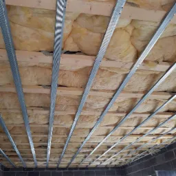 Ocieplenie stropu wełną mineralną pomiędzy drewnianymi belkami i metalowymi profilami, widok z dołu na surową konstrukcję przed montażem okładziny.