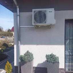 Biały klimatyzator marki AUX zamontowany na elewacji domu, obok rynny spustowej, z dwoma wysokimi donicami z iglakami u dołu i widokiem na ogród w tle.