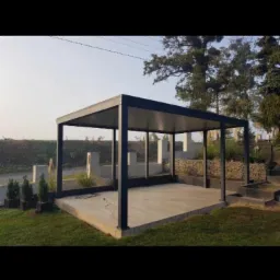 Nowoczesna, czarna pergola metalowa na betonowej platformie, otoczona zielenią i elementami dekoracyjnymi ogrodu, w słoneczny dzień.