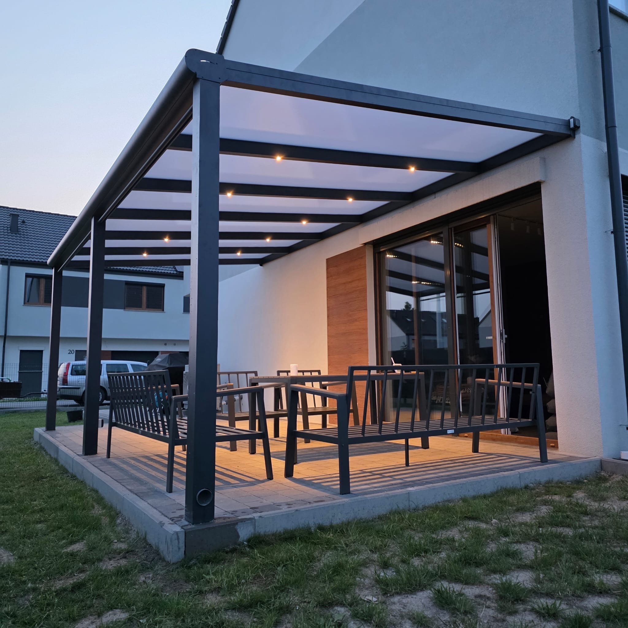 Elegancka, nowoczesna pergola metalowa z oświetleniem punktowym, tworząca zacienioną strefę relaksu z meblami ogrodowymi na tle domu.