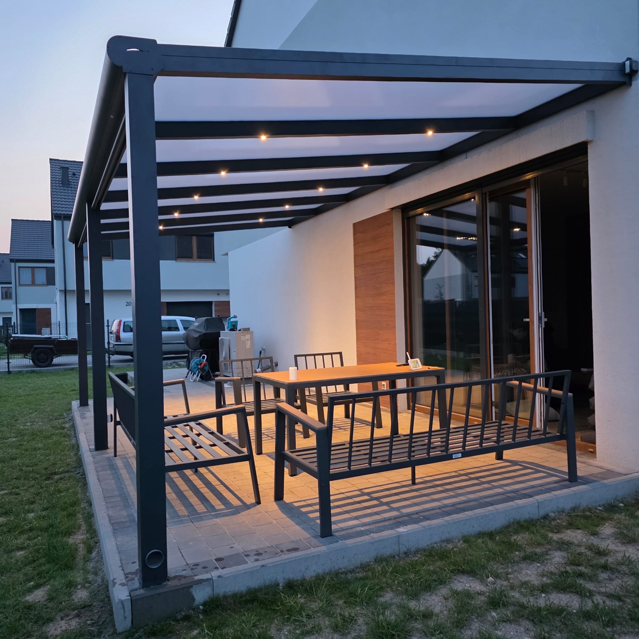 Nowoczesna, grafitowa pergola metalowa z oświetleniem LED, ustawiona na tarasie przy domu. Widoczne meble ogrodowe i przesuwne drzwi tarasowe.