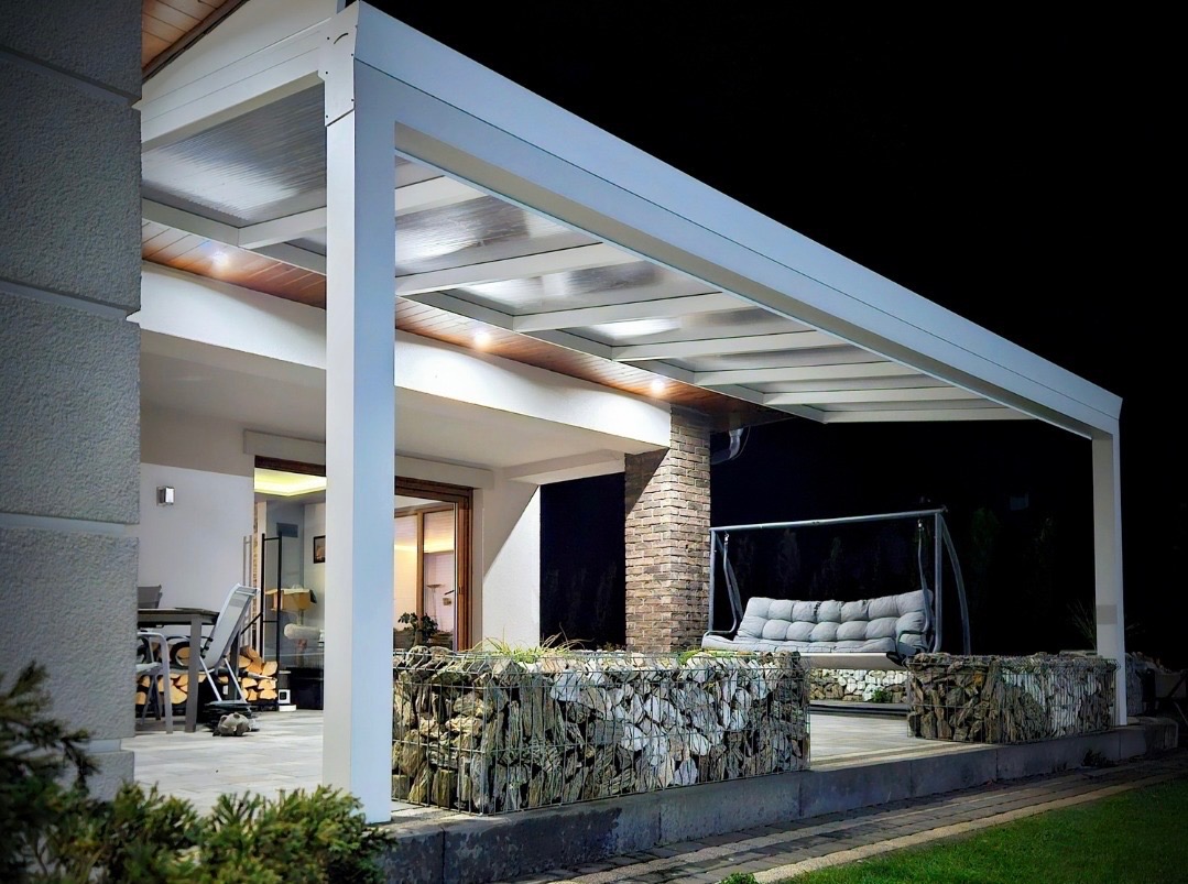 Biała pergola z transparentnym dachem, oświetlona, z gabionami i huśtawką w tle, nocne ujęcie.