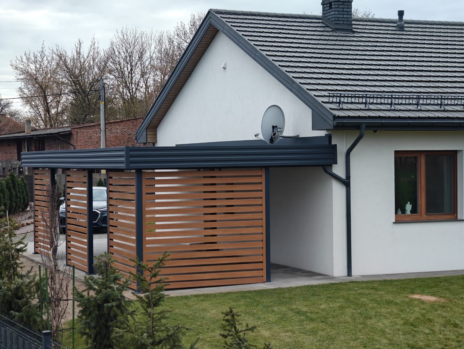 Nowoczesna, grafitowa pergola przy domu z drewnianymi panelami, tworząca zadaszone miejsce parkingowe. Widoczny fragment samochodu i zadbany trawnik.